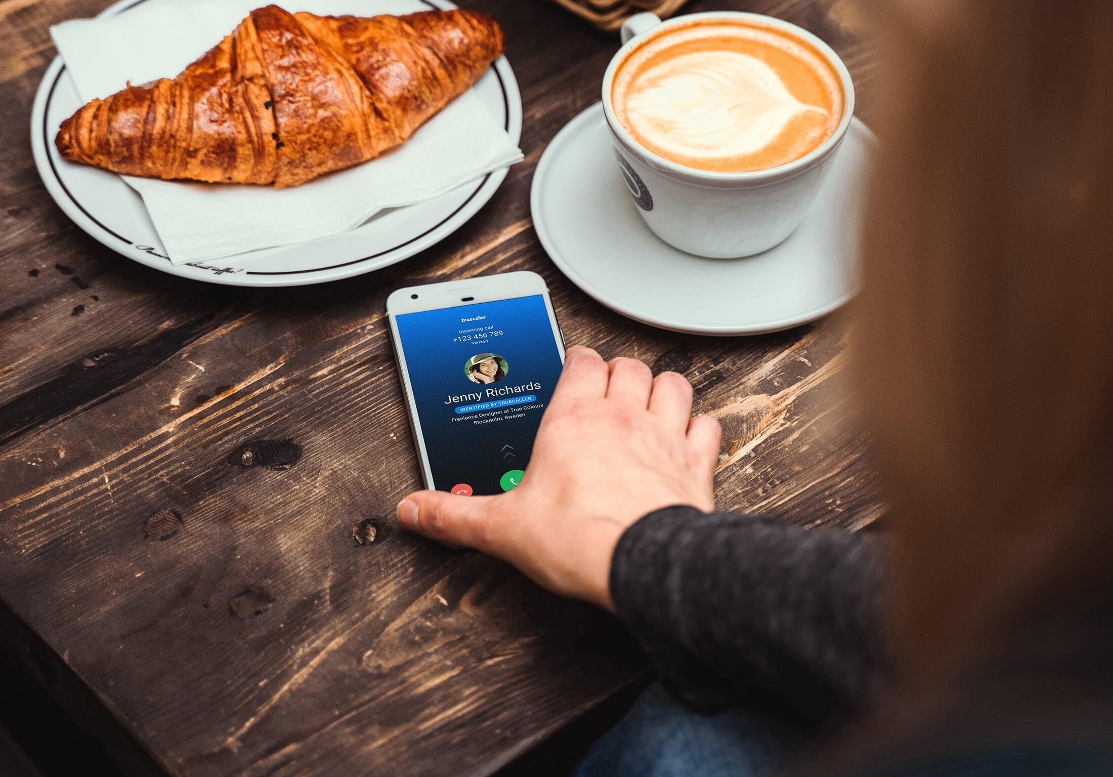 hand reaching for a phone on a coffee table, the phone is showing an incoming call by a "Jenny Richards" identified by Truecaller